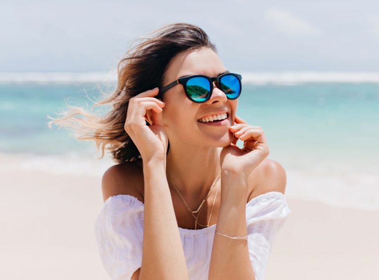 Wonderful woman in white attire and sparkle glasses posing with happy face expression in hot summer day. Pleasant caucasian girl standing near ocean on sky background.